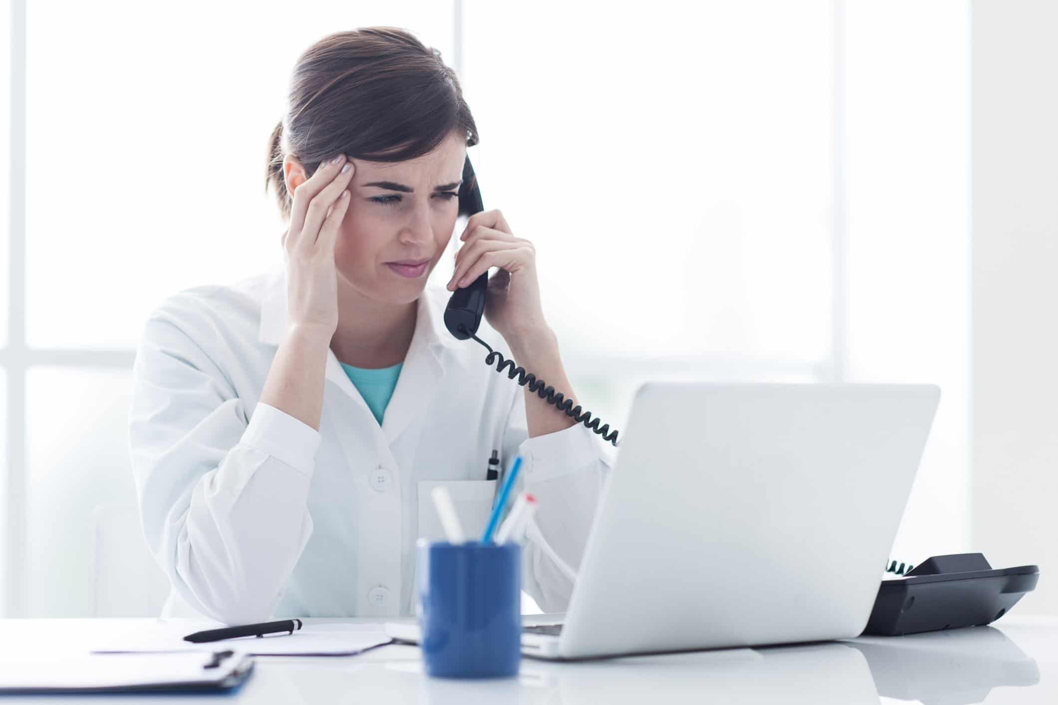 woman-sitting-at-desk-while-talking-on-corded-phone-and-holding-hand-at-temple-in-pain-from-migraine