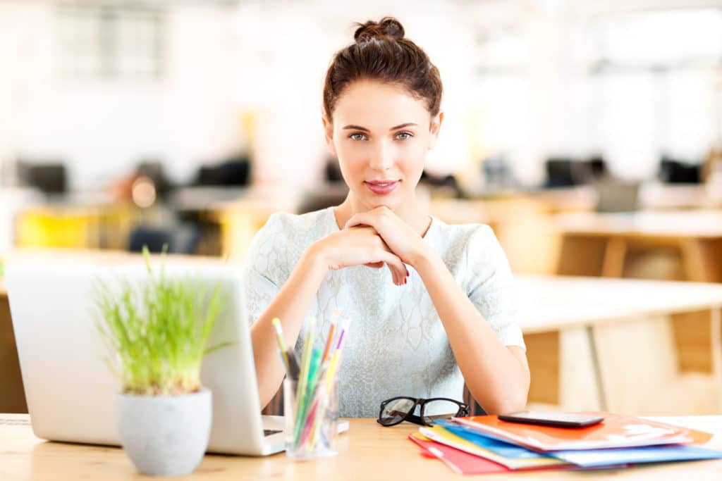 young-professional-woman-sitting-at-desk-with-hands-clasped-looking-at-camera-over-laptop-folders-and-potted-plant