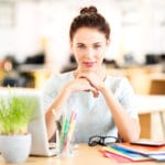 young-professional-woman-sitting-at-desk-with-hands-clasped-looking-at-camera-over-laptop-folders-and-potted-plant