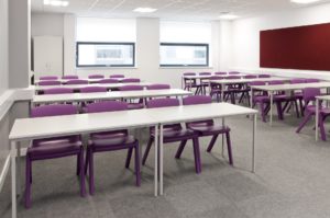 An empty classroom with rows of tables with purple chairs and overhead fluorescent lighting and two windows at the back of the room