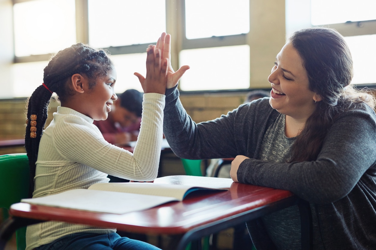 A woman teacher high fiving a young girl who is sitting at a single desk with an open book and windows with natural light in the background