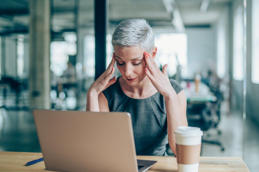 woman-putting-fingers-to-temples-while-sitting-in-office-with-laptop-and-coffee