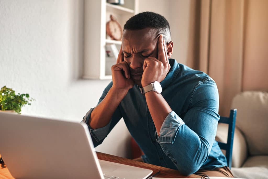 man-sitting-at-desk-with-hands-on-temples-with-migraine-and-laptop-on-desk