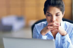 A businesswoman resting her chin on her clasped hands while looking at her laptop.