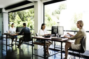 four-people-in-groups-of-two-working-at-tables-in-sunlit-office-next-to-large-windows