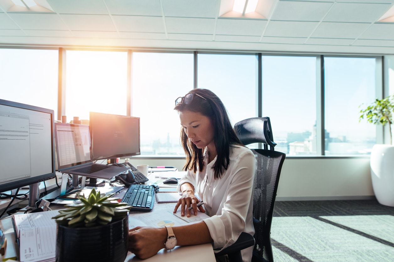 woman-sitting-at-desk-in-open-office-with-monitors-and-bright-windows