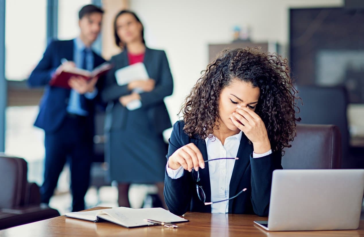 young-woman-holding-bridge-of-nose-while-sitting-at-office-desk-and-two-people-stand-in-background