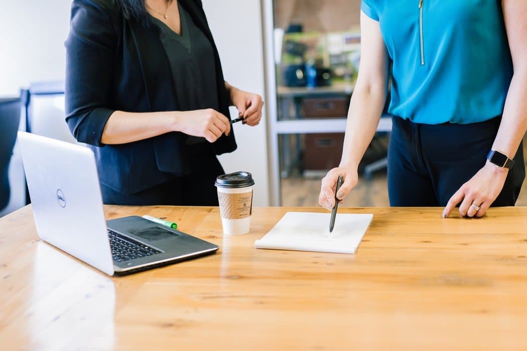 cropped-at-shoulders-photo-of-two-women-in-business-attire-standing-at-table-with-laptop-paper-pad-and-coffee-cup